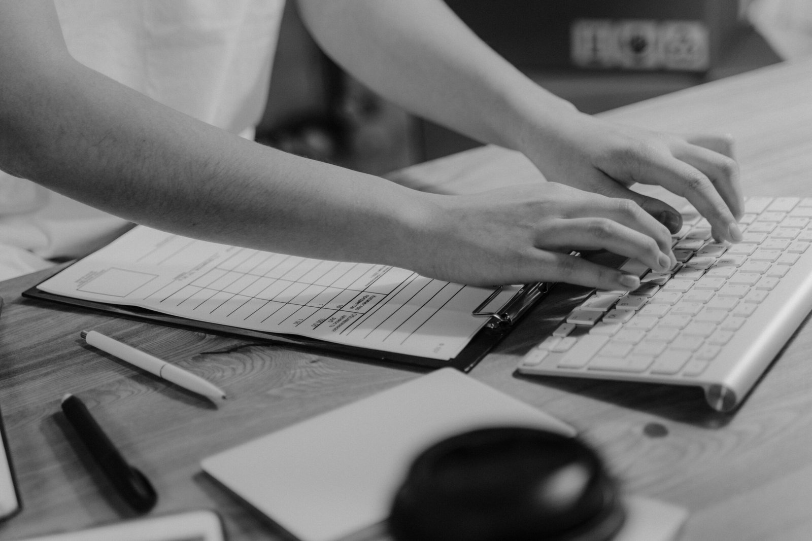 Black-and-white photo of hands typing on keyboard with clipboard on wooden desk.